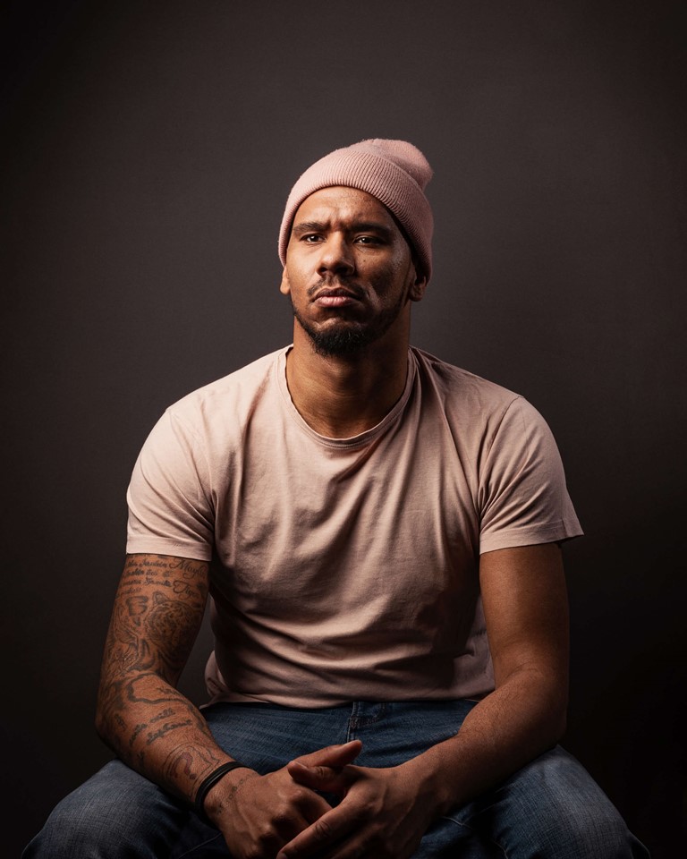 Sitting man of colour with beard and tattoos on his right arm. He is wearing a rose-coloured knitted cap, a bright T-shirt and blue jeans, his hands are intertwined.