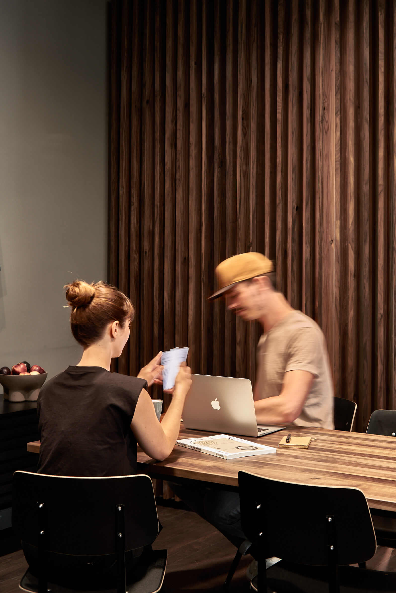 Ein junger Mann und eine Frau sitzen mit einem Laptop und weiterem Arbeitsmaterial an einem Holztisch, hinter den beiden befindet sich ein Sichtschutz aus dunklem Holz. 
