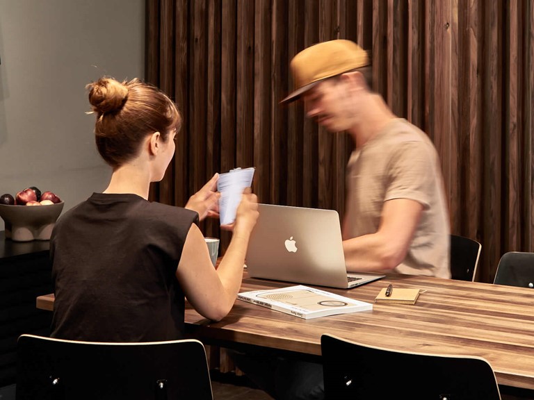 Ein junger Mann und eine Frau sitzen mit einem Laptop und weiterem Arbeitsmaterial an einem Holztisch, hinter den beiden befindet sich ein Sichtschutz aus dunklem Holz. 