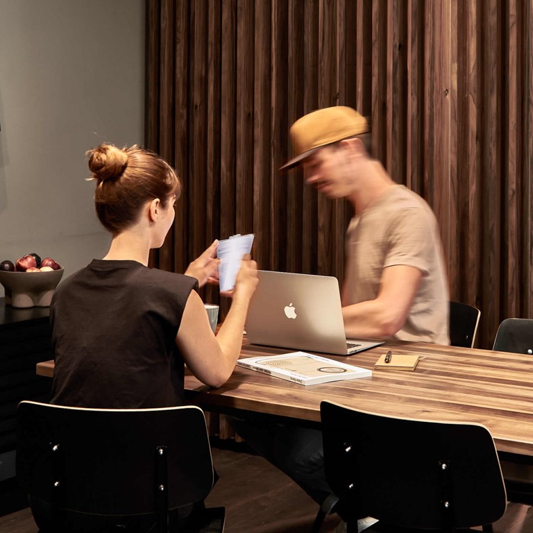 A young man and a woman sit at a wooden table with a laptop and other working materials, behind them is a dark wood screen. 