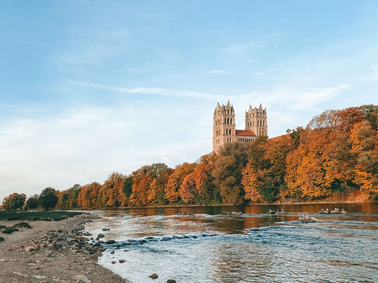 A riverbank is lined with autumn-coloured leafy trees, from which a church tower stands out, and there are birds on the water