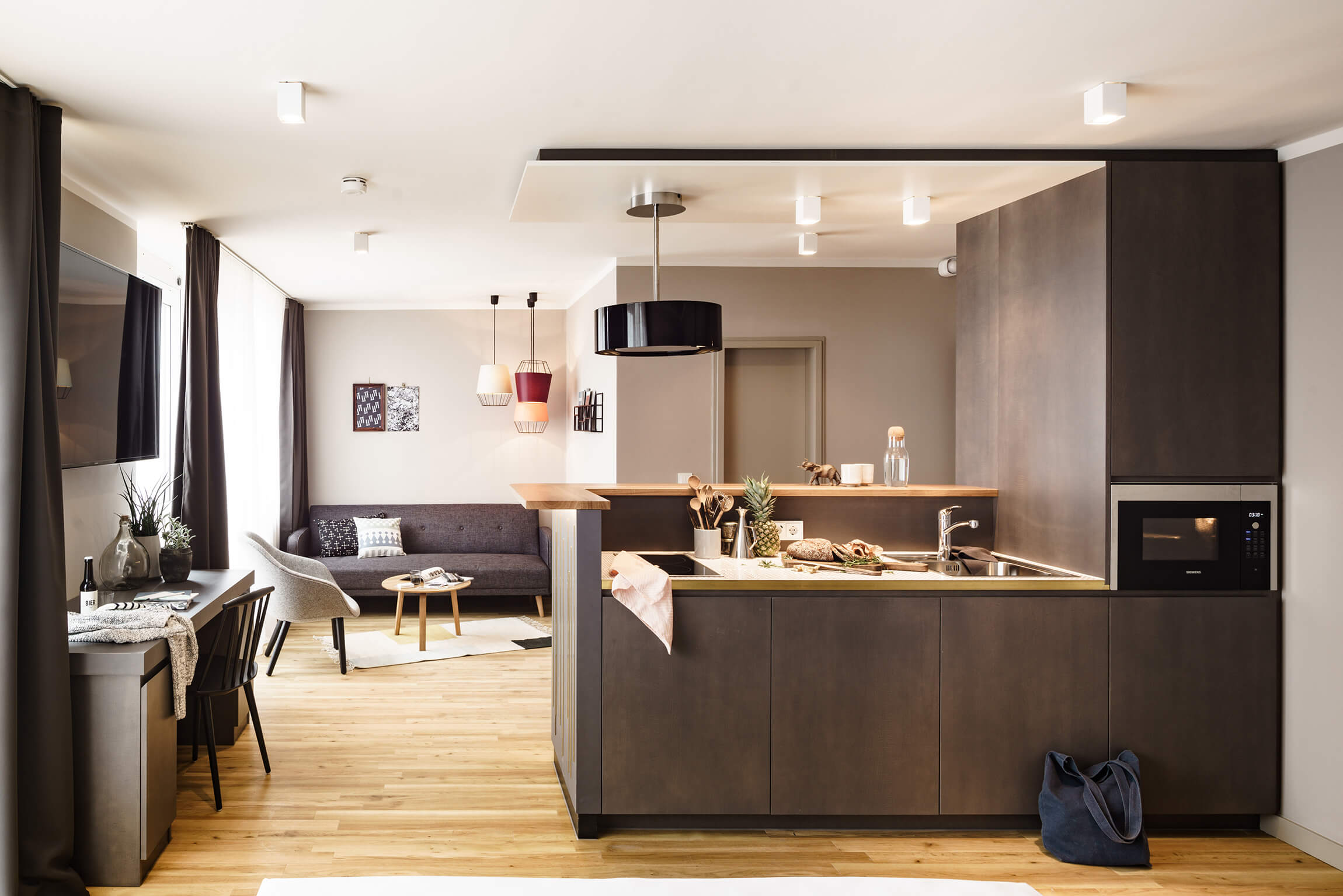 A bright flat with wooden flooring and a dark grey open-plan kitchen. On the left is a desk with accessories and above it a flat-screen TV. In the background, a large window with white curtains and a dark grey couch.