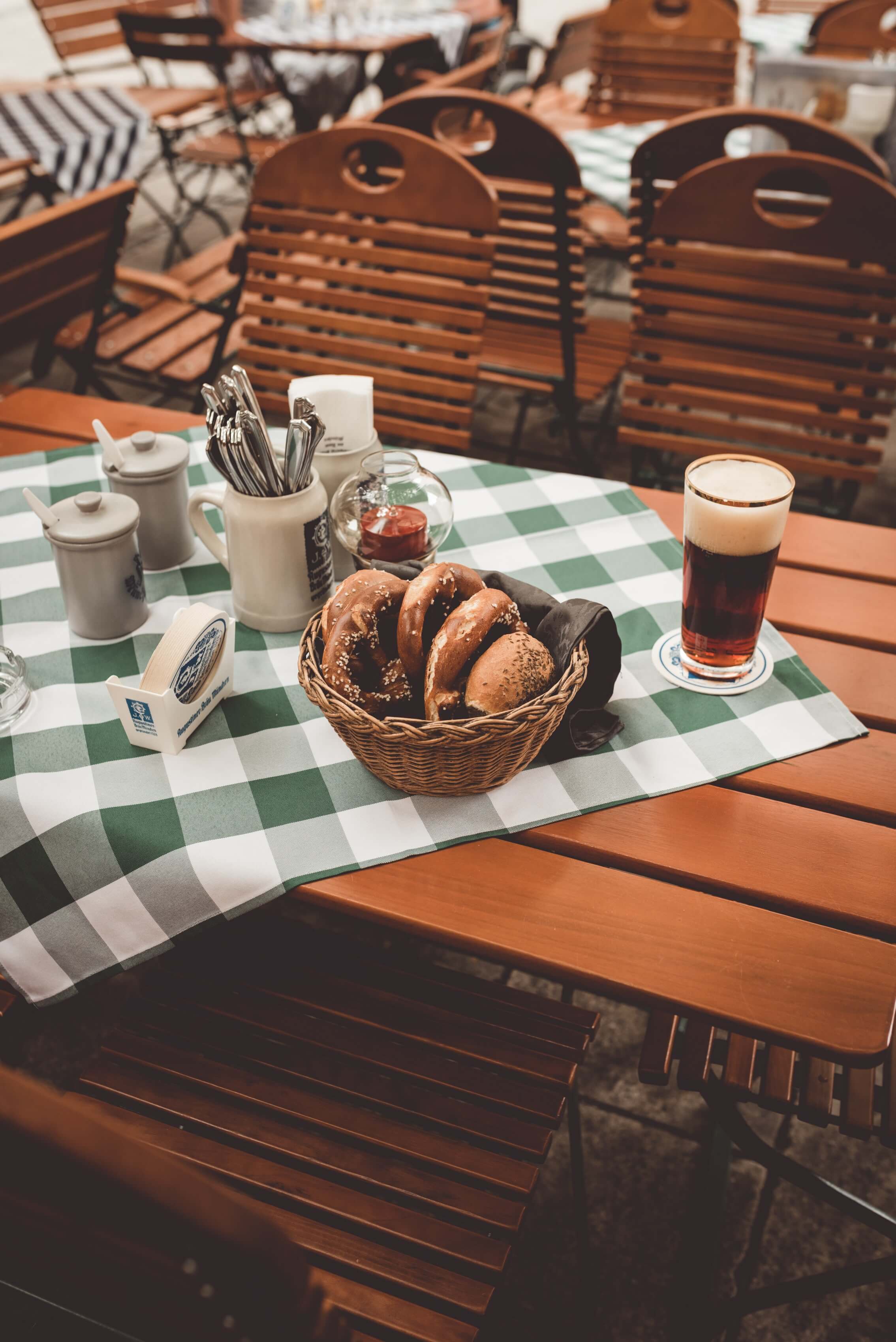Tables and chairs in a beer garden. On a green and white chequered tablecloth are a basket of pretzels, a glass of beer and cutlery
