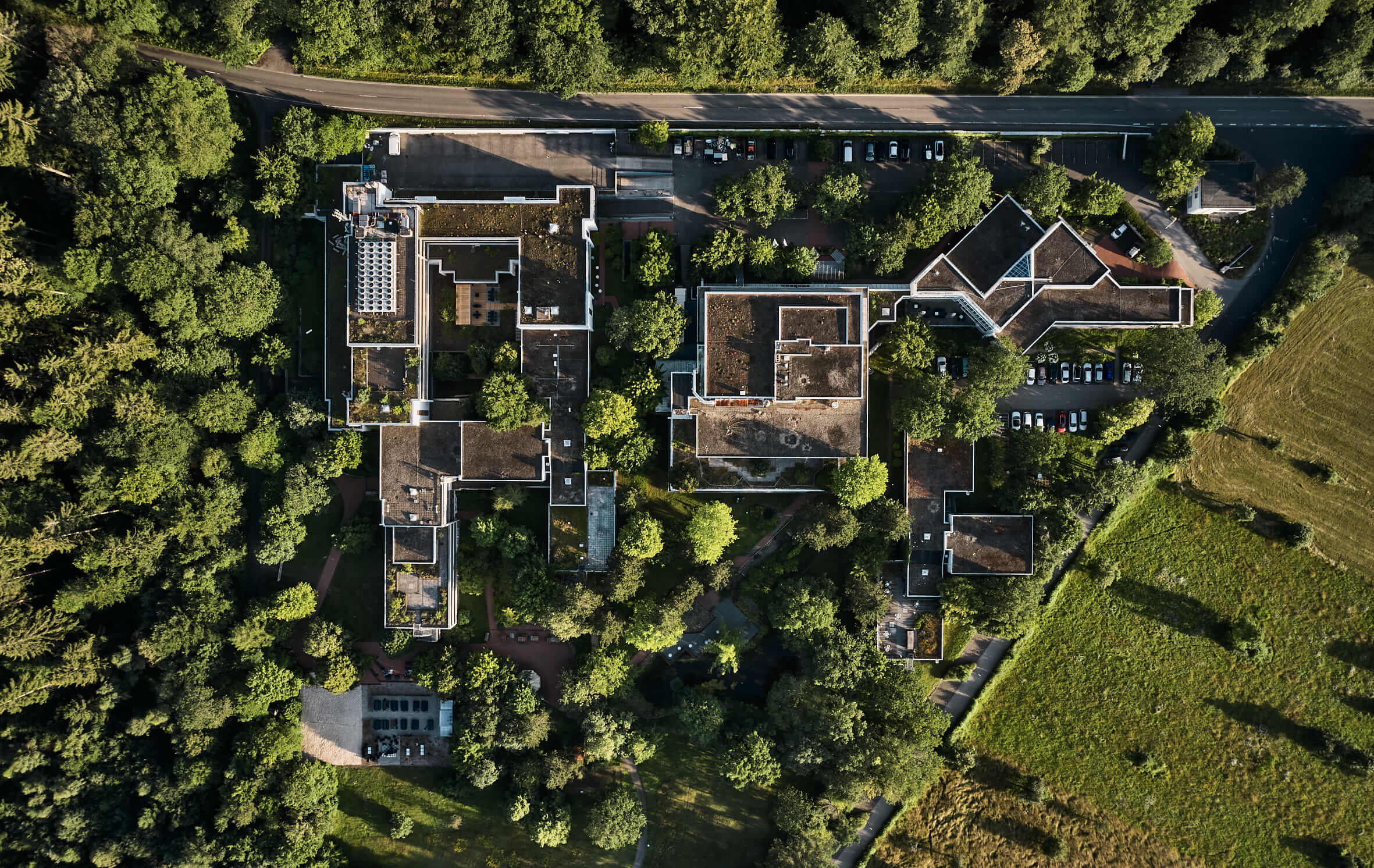 Ausgedehnter Gebäudekomplex der an einen Wald grenzt aus der Vogelperspektive, davor ein Park, rechts daneben ein Feld. Hinter den Gebäuden verläuft eine Straße.