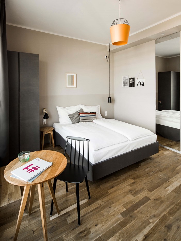 A white double bed in a room with a wooden floor, on the left a bedside table and a grey wardrobe, in front a round wooden table and black side chair.To the right of it a pendant lamp and a large wall mirror.