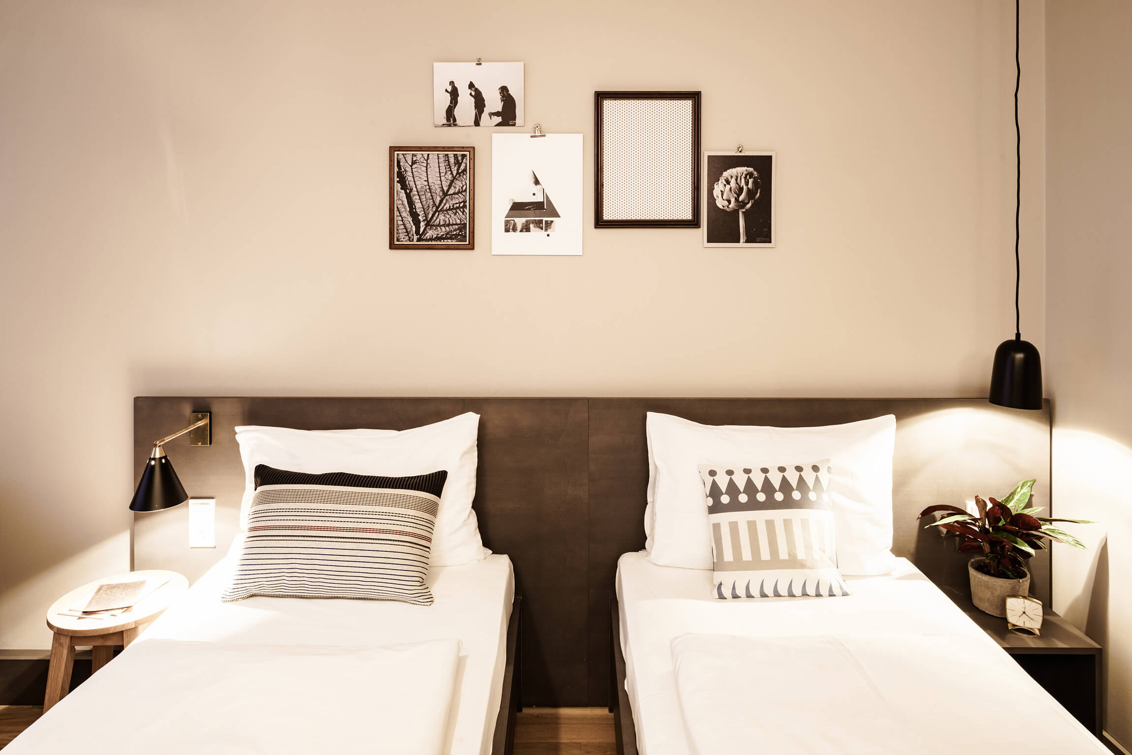 Zwei gemütlich aussehende Einzelbetten neben denen je ein Nachttisch mit Wasserglas, Buch, Pflanze stehen, darüber sind Leuchten angebracht. Die Wand ist mit schwarz-weißen Bildern dekoriert.