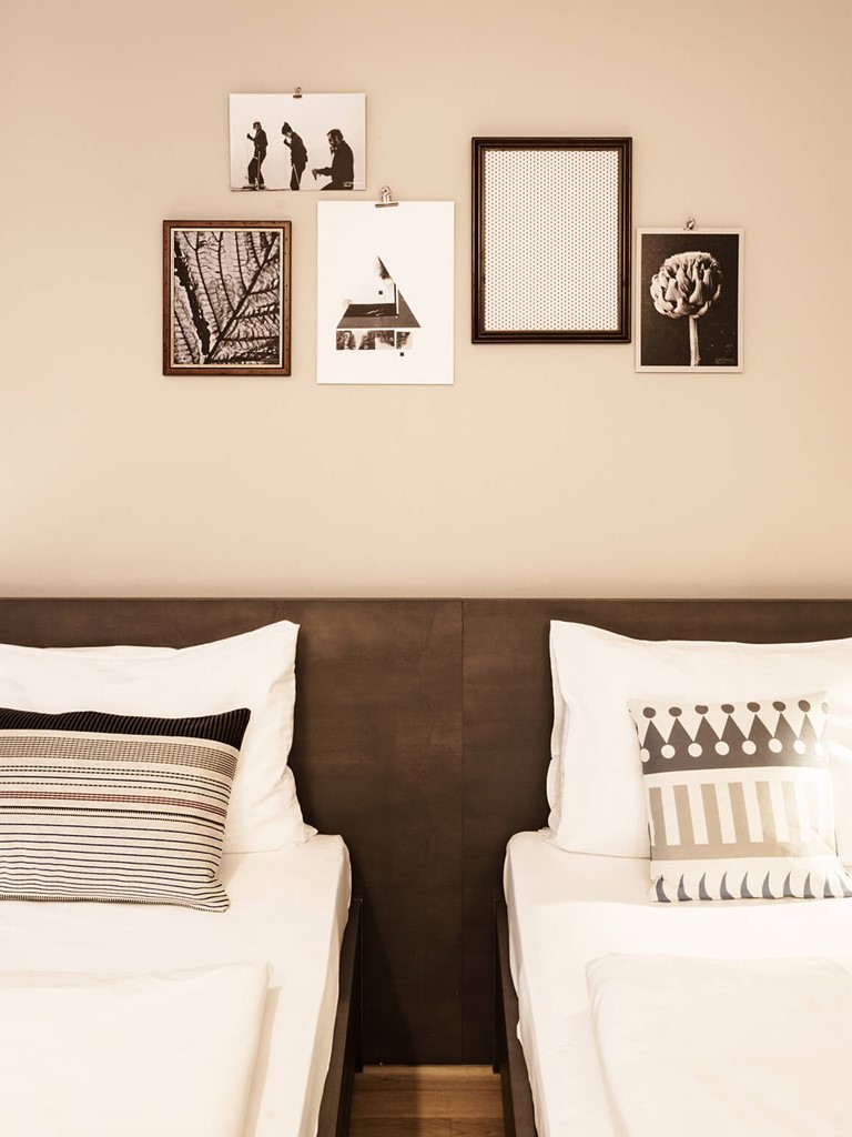 Two cosy-looking single beds, each with a bedside table with a glass of water, or a notebook and a plant, and lamps above them. The wall is decorated with black and white pictures.