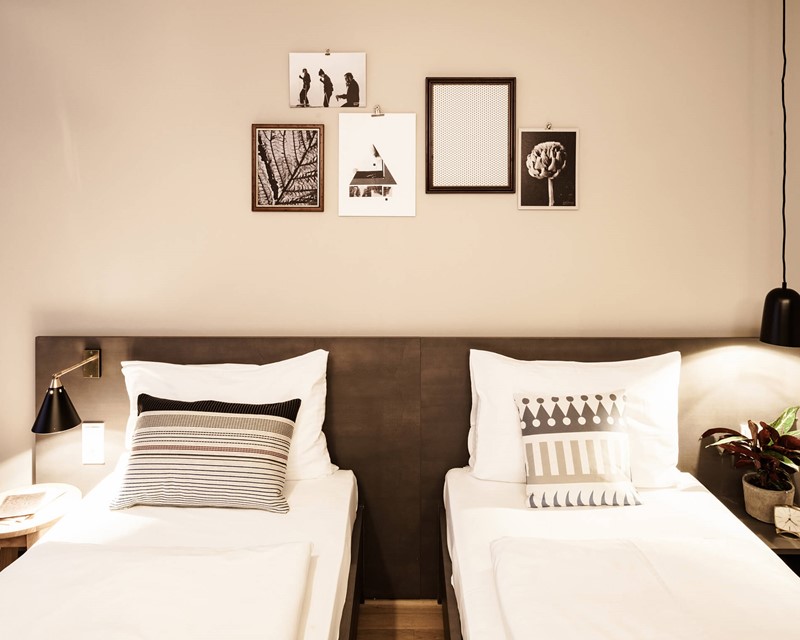 Two cosy-looking single beds, each with a bedside table with a glass of water, or a notebook and a plant, and lamps above them. The wall is decorated with black and white pictures.