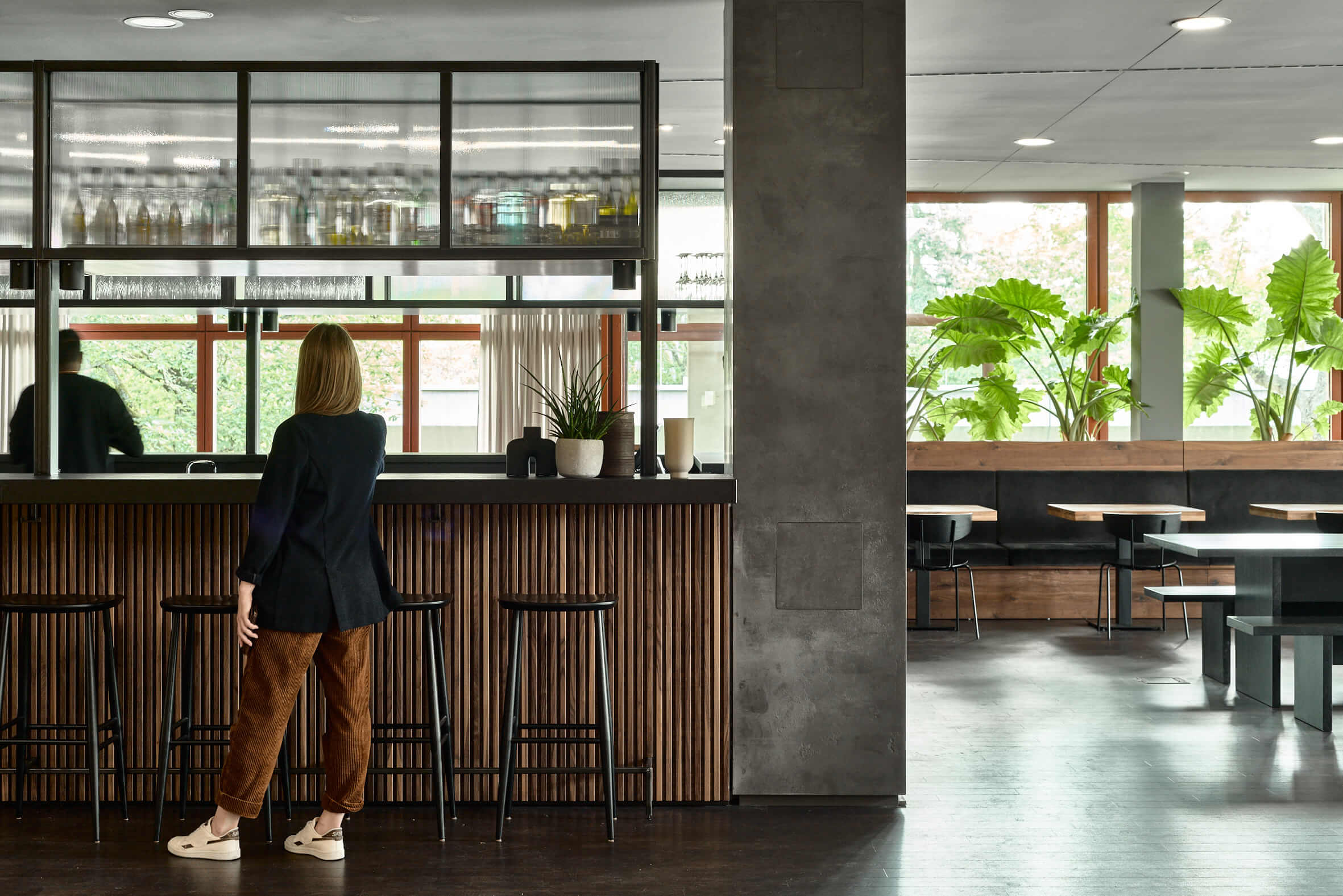 Eine moderne Bar mit Barhockern an der eine Frau steht, rechts sind Tische, Stühle und Sitzbänke zu sehen sowie große Fensterfronten mit Blick ins Grüne. Rechts sind Tische, Sitzbänke und Stühle zu sehen, im Hintergrund eine Fensterfront mit Grünpflanzen.