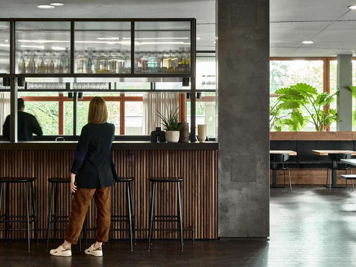 A modern bar with bar stools at which a woman is standing, on the right are tables, benches and chairs, in the background a window front with green plants.
