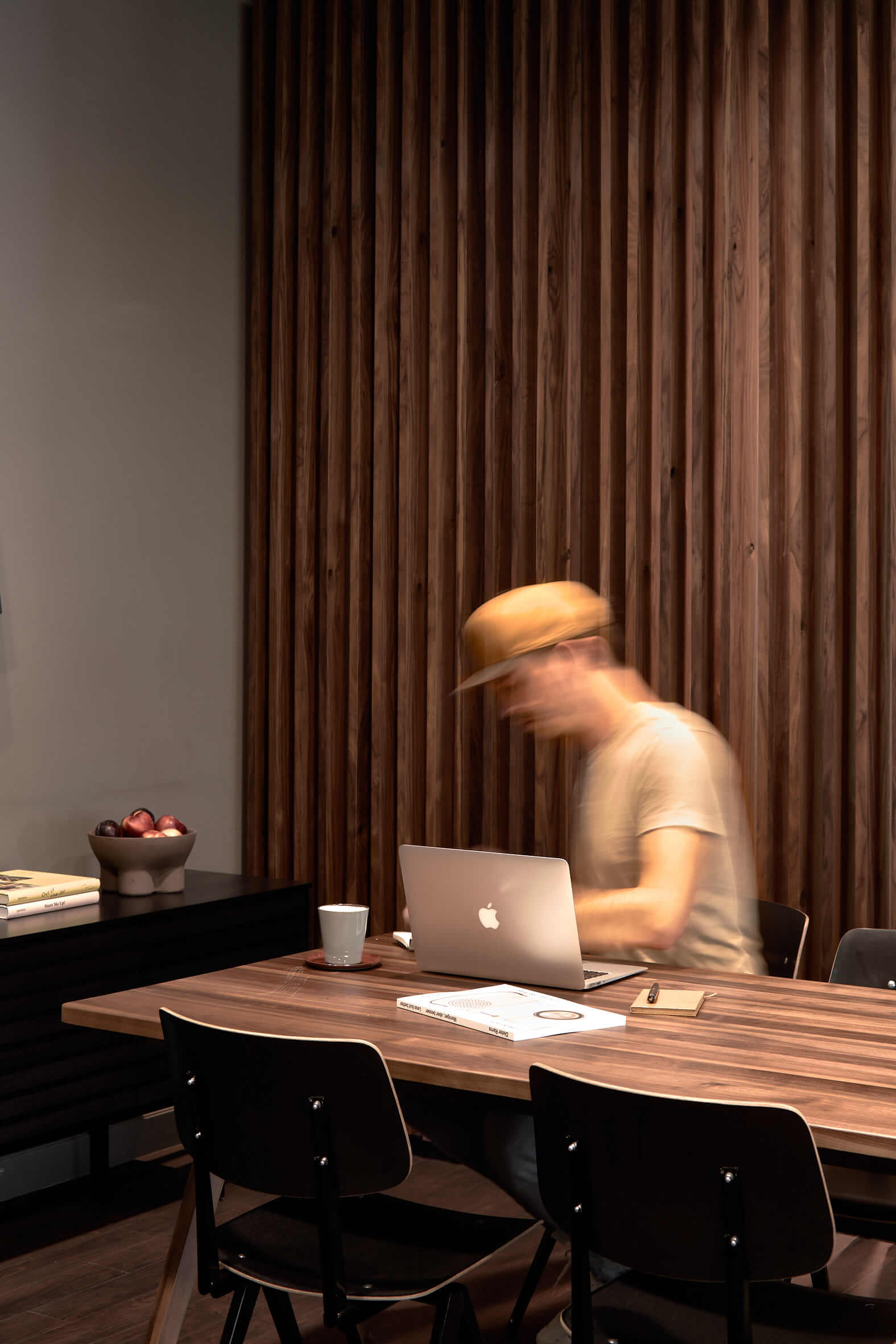Großer Holztisch an dem schwarze Stühle stehen, ein Mann mit gelber Basecap sitzt an einem MacBook und arbeitet. Links davon ein dunkles Sideboard mit einer Obstschale.