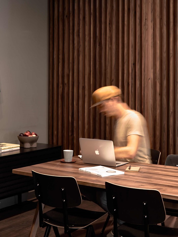 Large wooden table with black chairs, a man wearing a yellow baseball cap sitting at a MacBook and working. To the left is a dark sideboard with a fruit bowl.