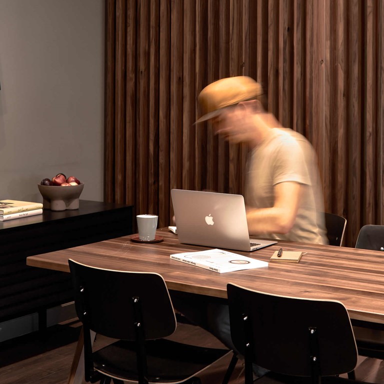 Large wooden table with black chairs, a man wearing a yellow baseball cap sitting at a MacBook and working. To the left is a dark sideboard with a fruit bowl.