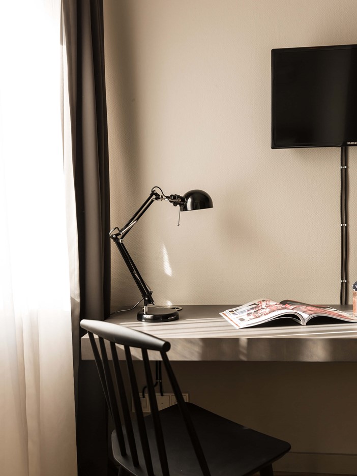Anschnitt eines Fensters mit Vorhängen und einem Schreibtisch auf dem sich eine schwarze Lampe und ein Magazin befinden, davor ein schwarzer Stuhl. Ein wandmontierter Flachbildfernseher ist zu erkennen.