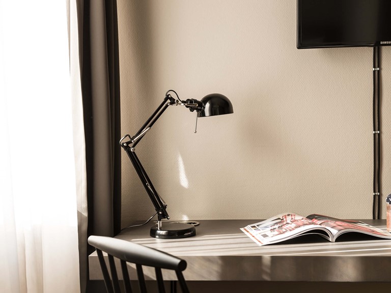 Detail of a window with curtains and a desk with a black lamp and a magazine on it, a black chair in front of it. There is also a flat-screen TV mounted on the wall.