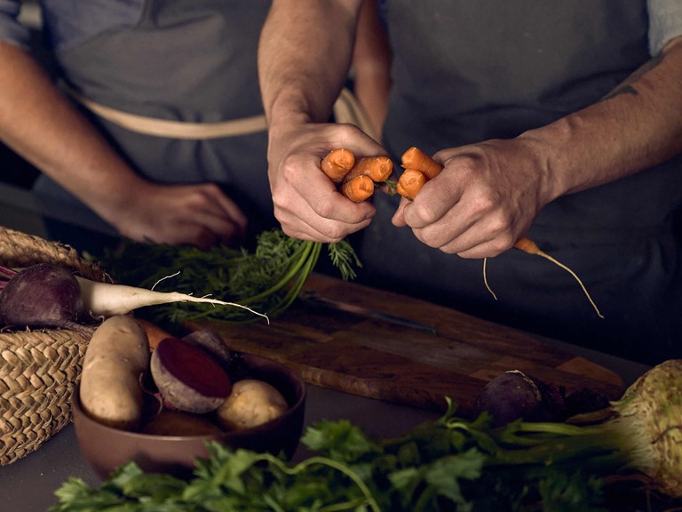 Root vegetables on a wooden table, above it the arms of a person who is snapping a small bunch of carrots in half