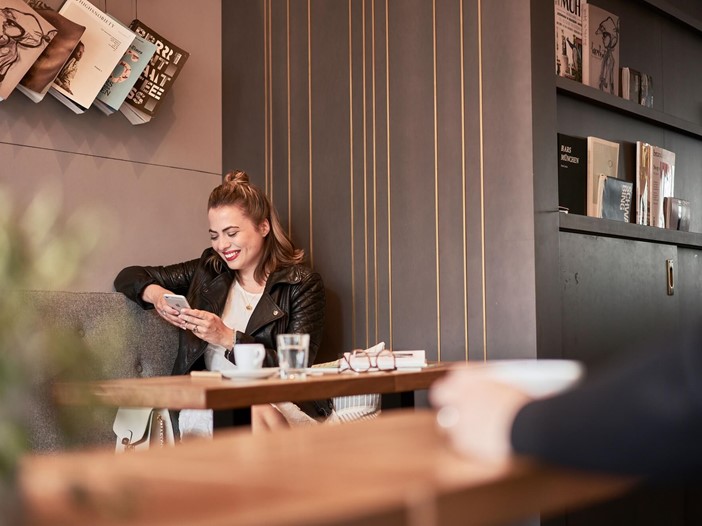 Eine braunhaarige, junge Frau sitzt lächelnd in der Sitzecke einer Lobby. Sie trägt blickt auf ihr Smartphone. Vor ihr auf dem Tisch stehen Kaffee und ein Glas Wasser, Brille und Buch liegen daneben.