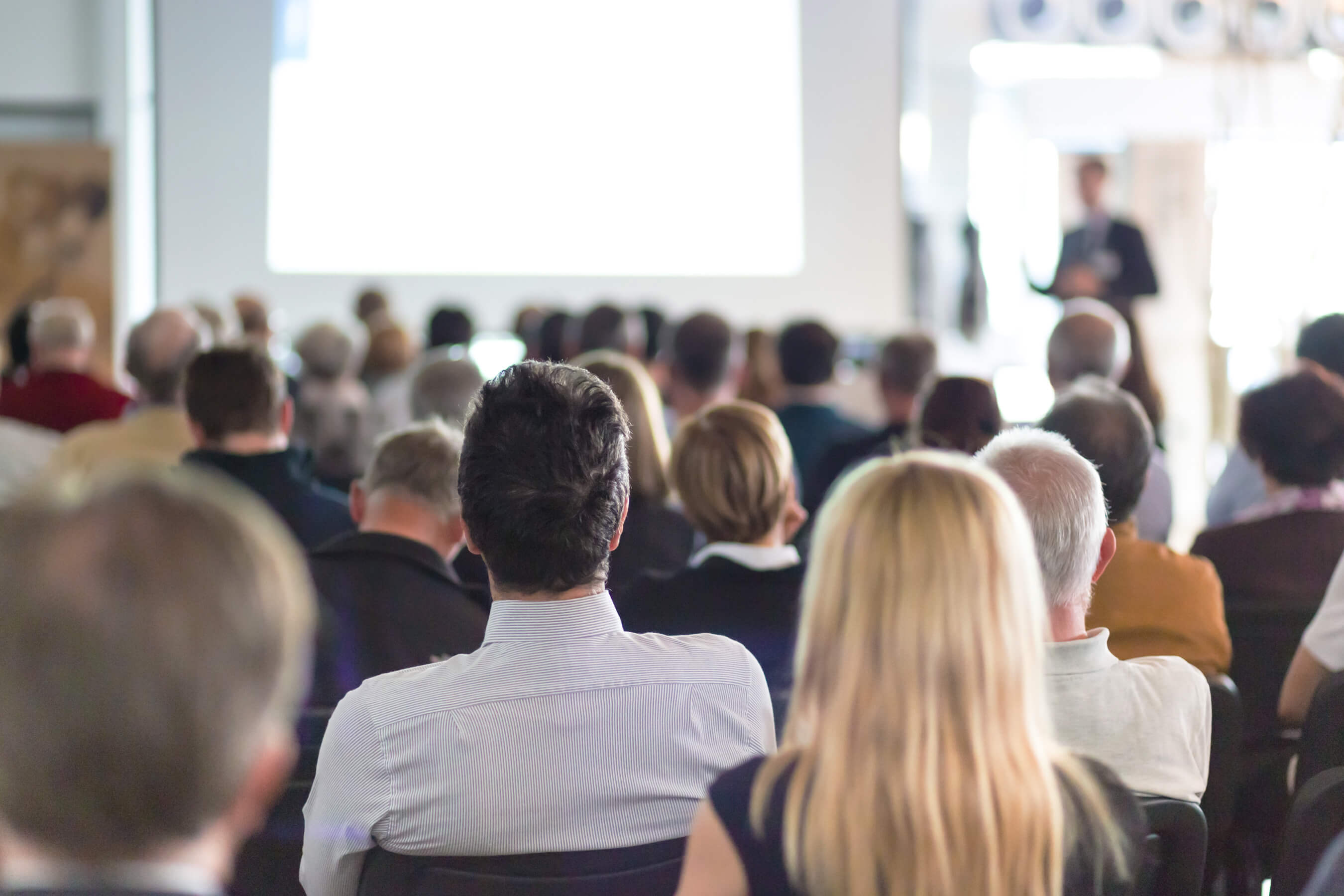 Geschäftsmäßig aussehende Personen in einem großen Raum. Vor den Personen im Hintergrund zu erkennen, ist eine Leinwand und Mann im Anzug hält eine Präsentation.