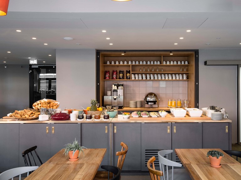 A counter with a rich breakfast selection, behind it a coffee bar. In front of the counter are chairs of different colours at wooden tables.