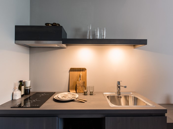 Detail of an illuminated, dark grey kitchenette, on it various accessories such as wooden boards, glasses and carafes. 