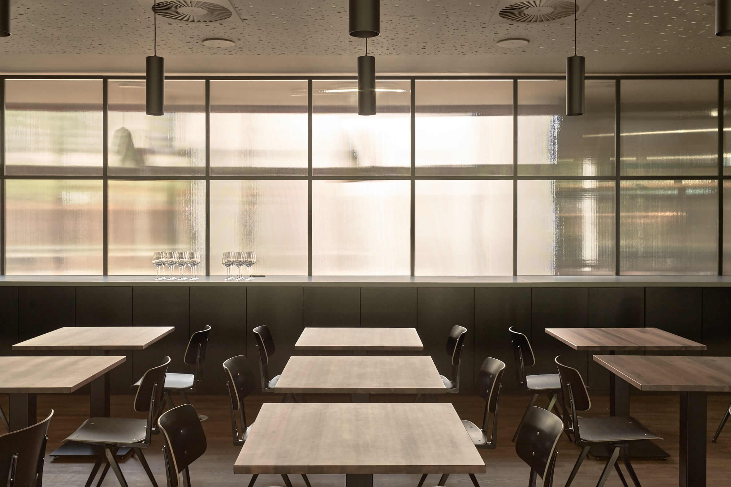 Restaurant mit dunklen Stühlen an kleineren Holztischen. Den Hintergrund bestimmt eine Milchglaswand, die viel Licht herein lässt.