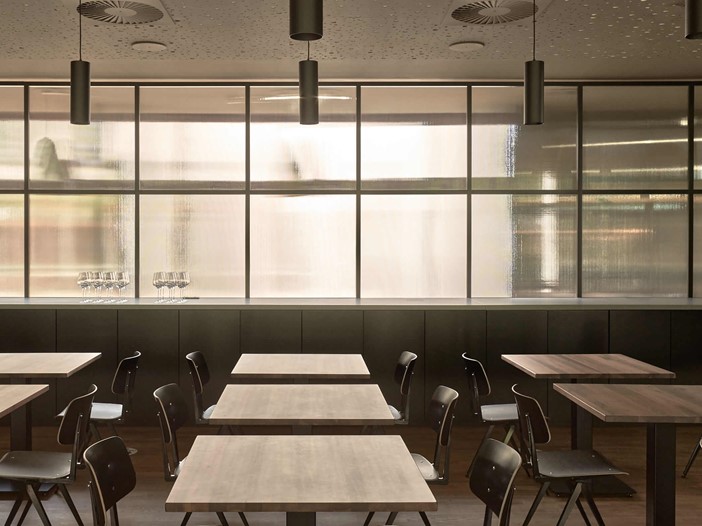 Restaurant mit dunklen Stühlen an kleineren Holztischen. Den Hintergrund bestimmt eine Milchglaswand, die viel Licht herein lässt.