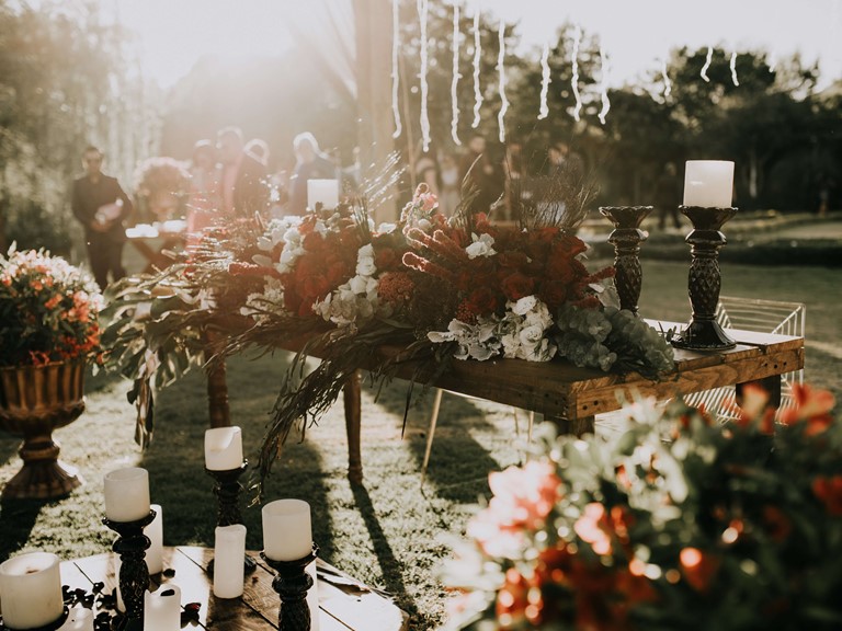 An outdoor table decorated with red and white floral arrangements and lots of white candles. Behind it you can see smartly dressed people.
