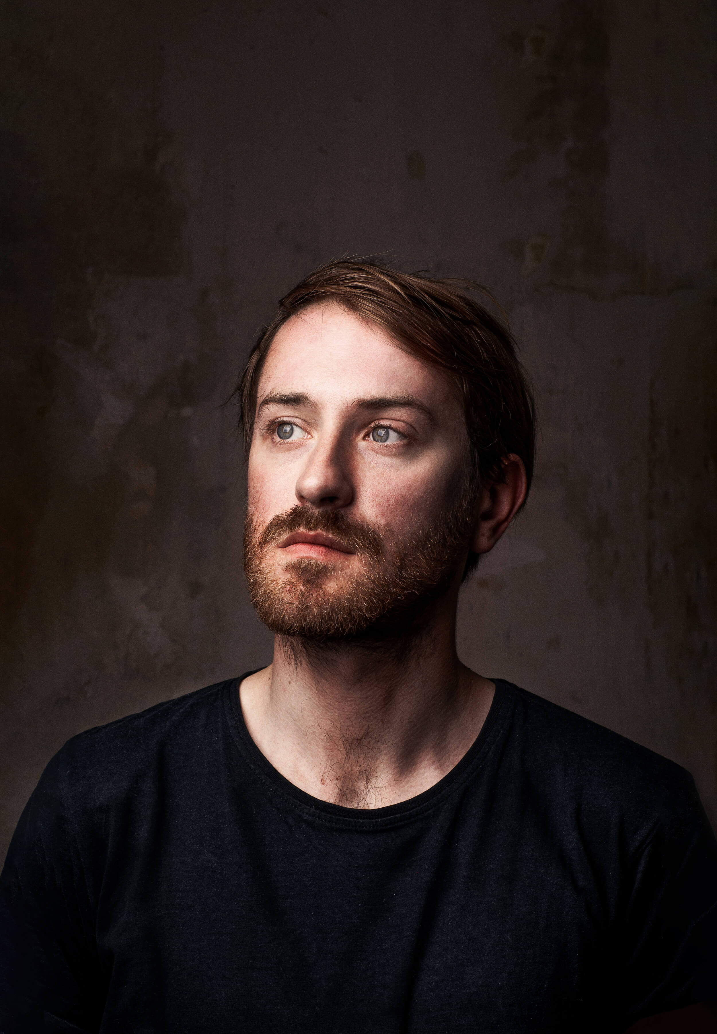 Portrait eines rothaarigen Mannes mit Vollbart vor einem dunkelgrauen Hintergrund. Er trägt ein schwarzes Oberteil und blickt nach oben rechts ins Leere.
