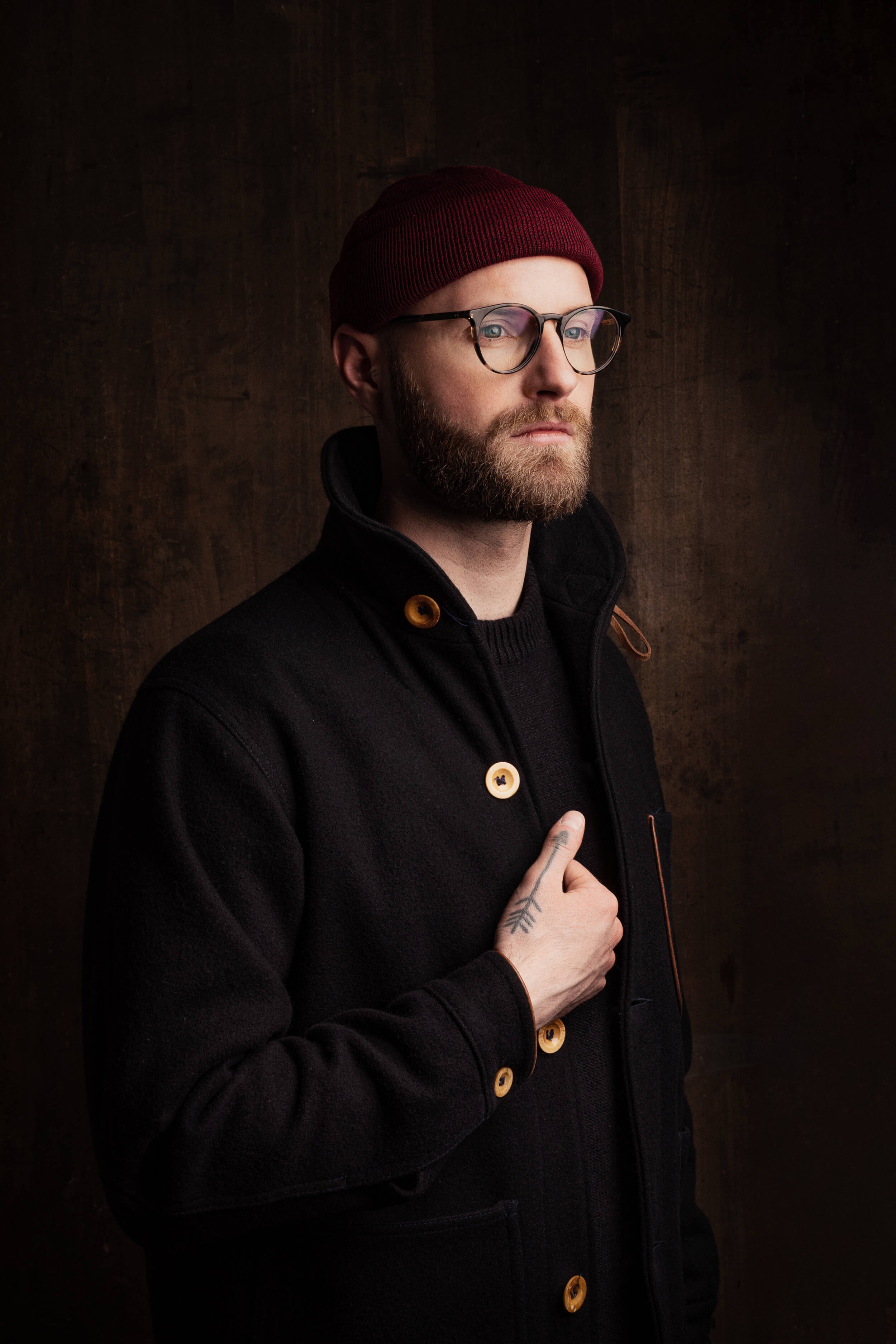 Mann mit Vollbart und Hornbrille. Er trägt eine bordeauxfarbene Strickmütze und eine schwarze Jacke mit großen hölzernen Knöpfen. Die rechte Hand am Revers ziert ein Tattoo in Pfeil-Form.