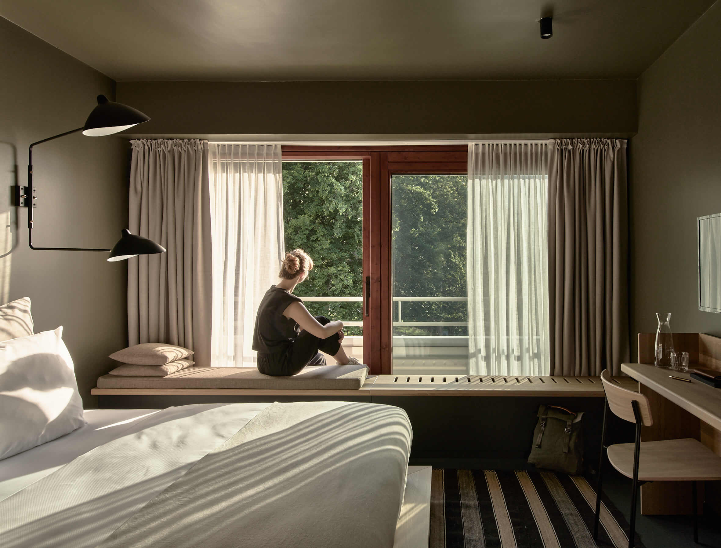 Junge Frau sitzt auf einer Sitzbank in einem modernen, in erdenden Tönen gehaltenen Hotelzimmer und blickt aus dem Fenster ins Grüne. 