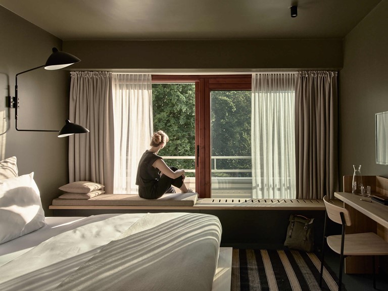 A woman sits on a bench in a modern hotel room decorated in earthy tones and looks out of the window into the greenery. 