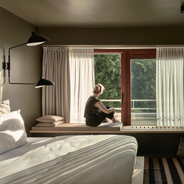 A woman sits on a bench in a modern hotel room decorated in earthy tones and looks out of the window into the greenery. 