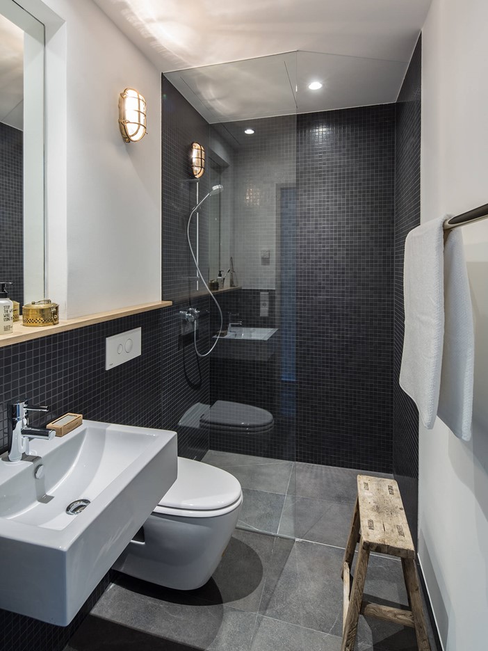 Modernes, grau gefliestes Badezimmer, links ein weißes Waschbecken und WC, am Ende des Raumes eine begehbare Dusche mit Glastür.