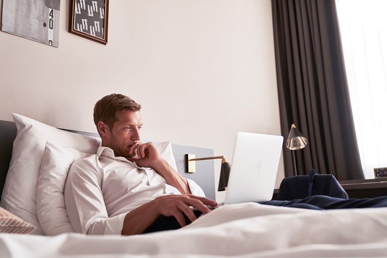 Man in white shirt is lying on a bed with his notebook on his lap, looking contemplative