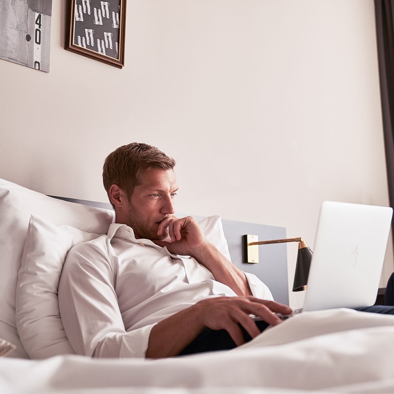 Mann in weißem Hemd liegt auf einem Bett und hat seinen Laptop auf dem Schoß, er wirkt nachdenklich