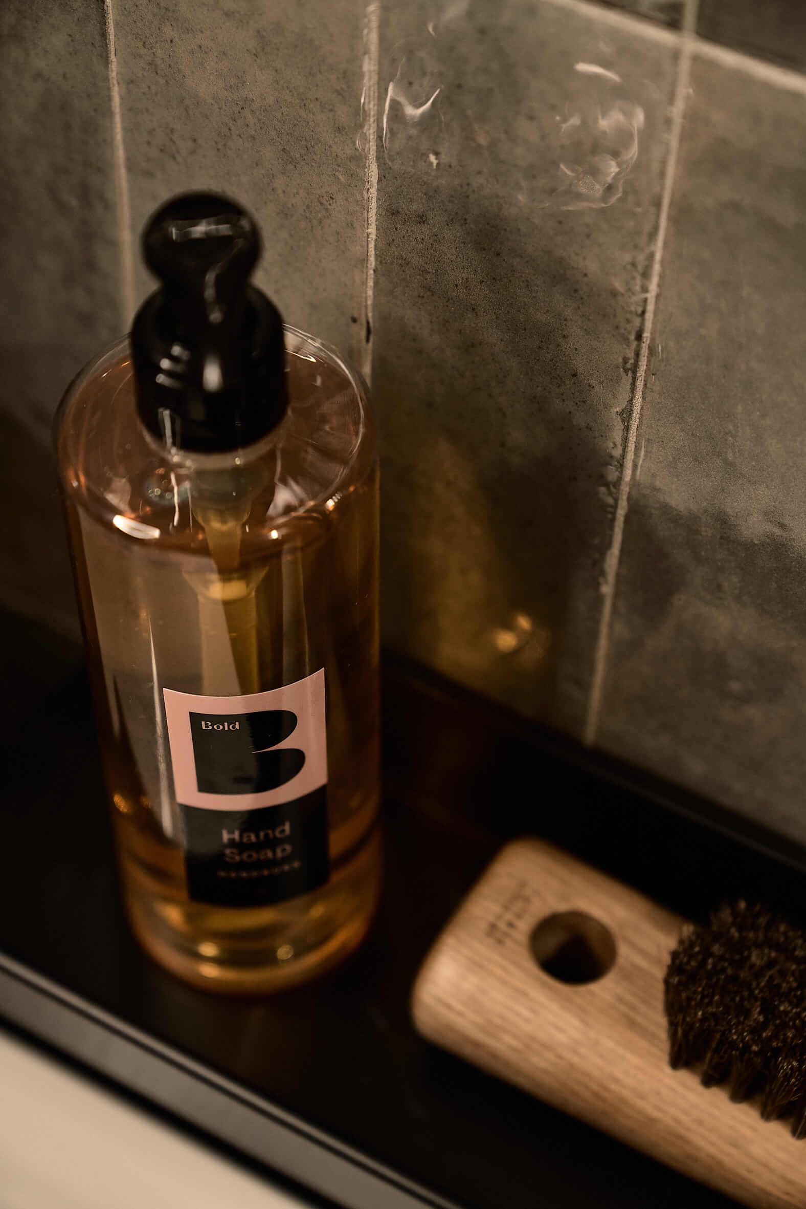 Transparent soap dispenser with sticker and gold-coloured contents on a dark shelf, next to it a wooden brush with dark bristles.