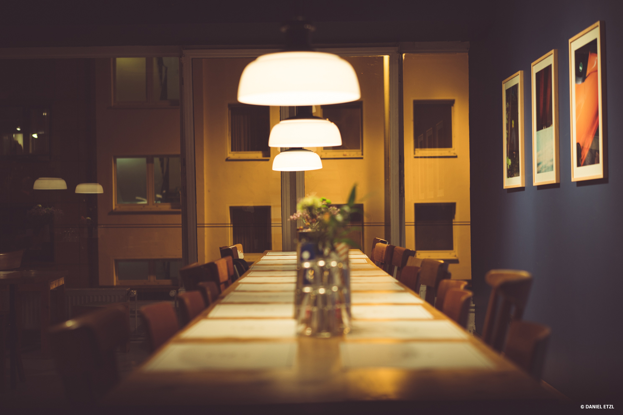 Ein langer Tisch mit verschiedenen Holzstühlen, rechts an der blauen Wand mehrere Bilder, alles erleuchtet durch drei Hängelampen über dem Tisch
