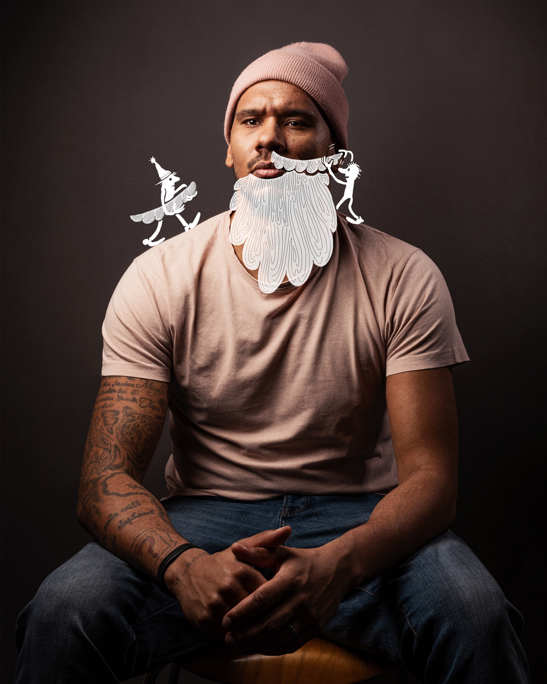  Sitzender Mann of Colour mit Bart, Tattoos und einer Strickmütze, seine Hände sind ineinander verschränkt, zwei illustrierte Männchen kleben ihm einen Vollbart an