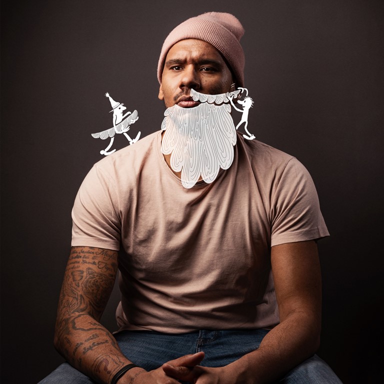  Sitting man of colour with beard, tattoos and a knitted beanie, his hands are intertwined, two illustrated figures stick a full beard on him