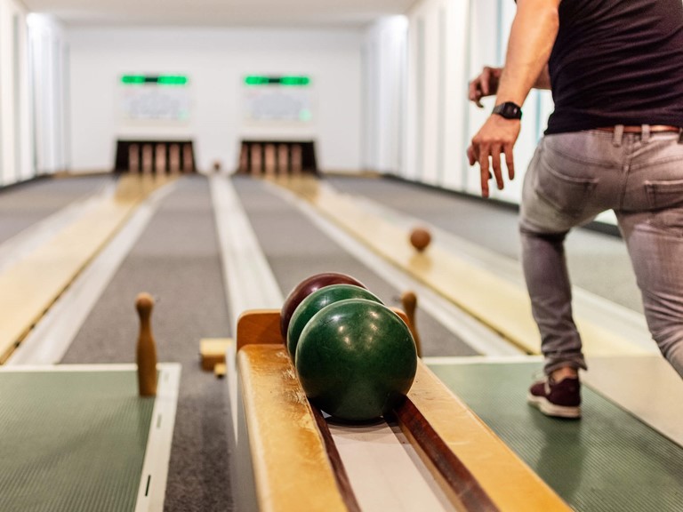 A man in grey jeans and a black T-shirt playing skittles. The ball rolls on the right lane towards the pins