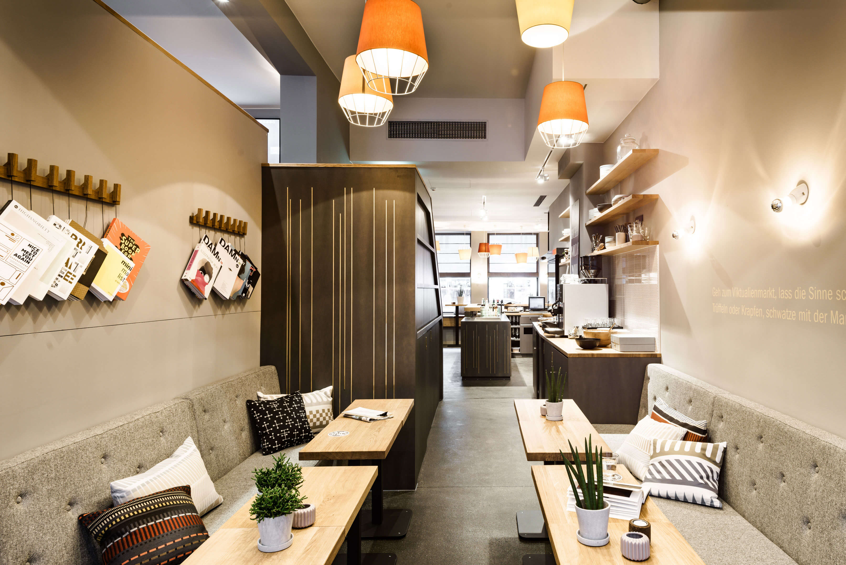 Lounge overlooking a bar and tables with chairs at the back of the window. Upholstered benches with wooden tables and colourful decorative cushions and accessories. Books hang on hooks on the wall to the left. 