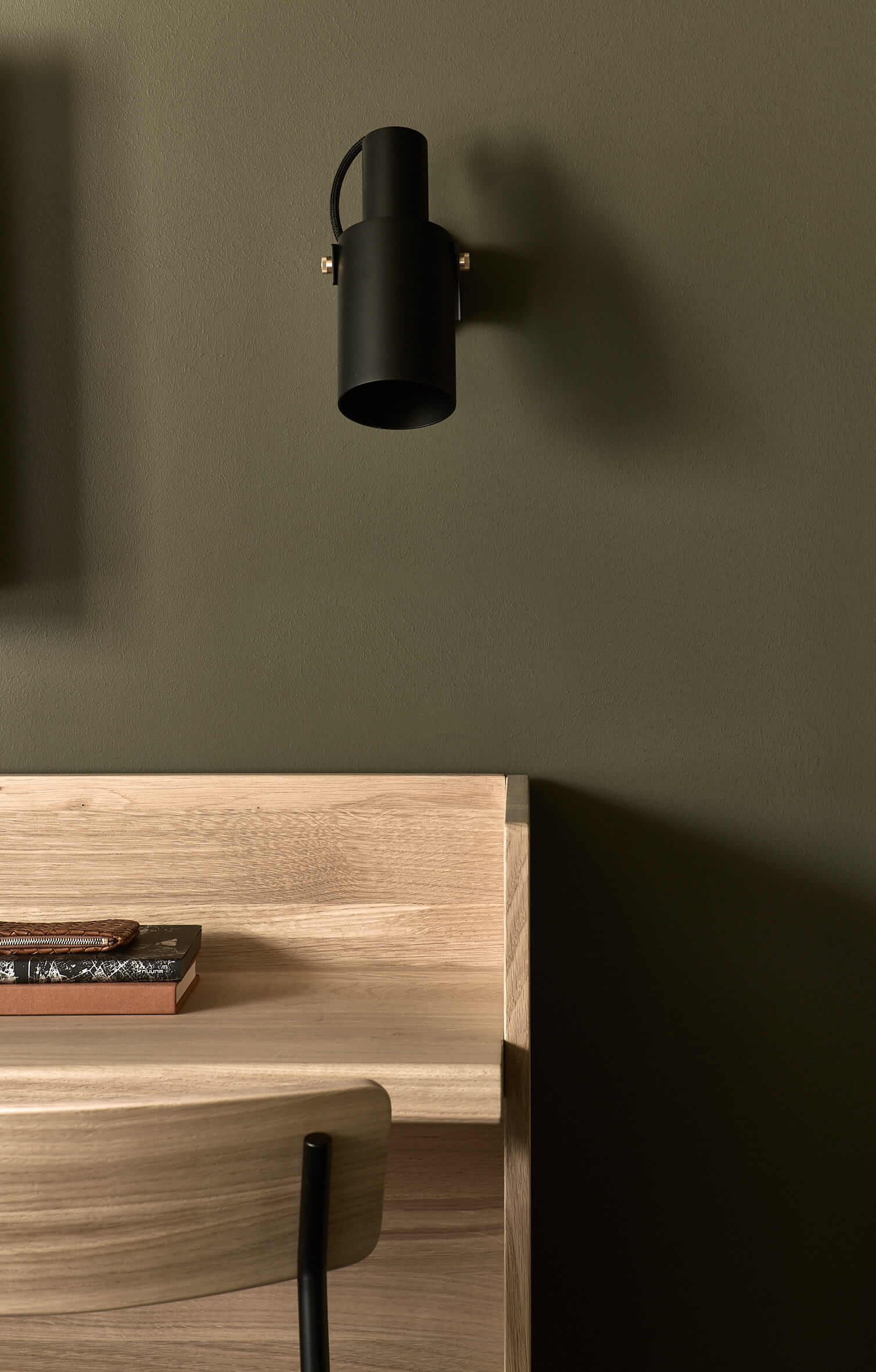 Close-up of a light, wooden desk against a dark green wall with a chair in front of it, above it a small black wall lamp. 
