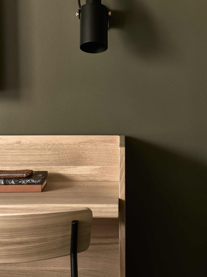 Close-up of a light, wooden desk against a dark green wall with a chair in front of it, above it a small black wall lamp. 