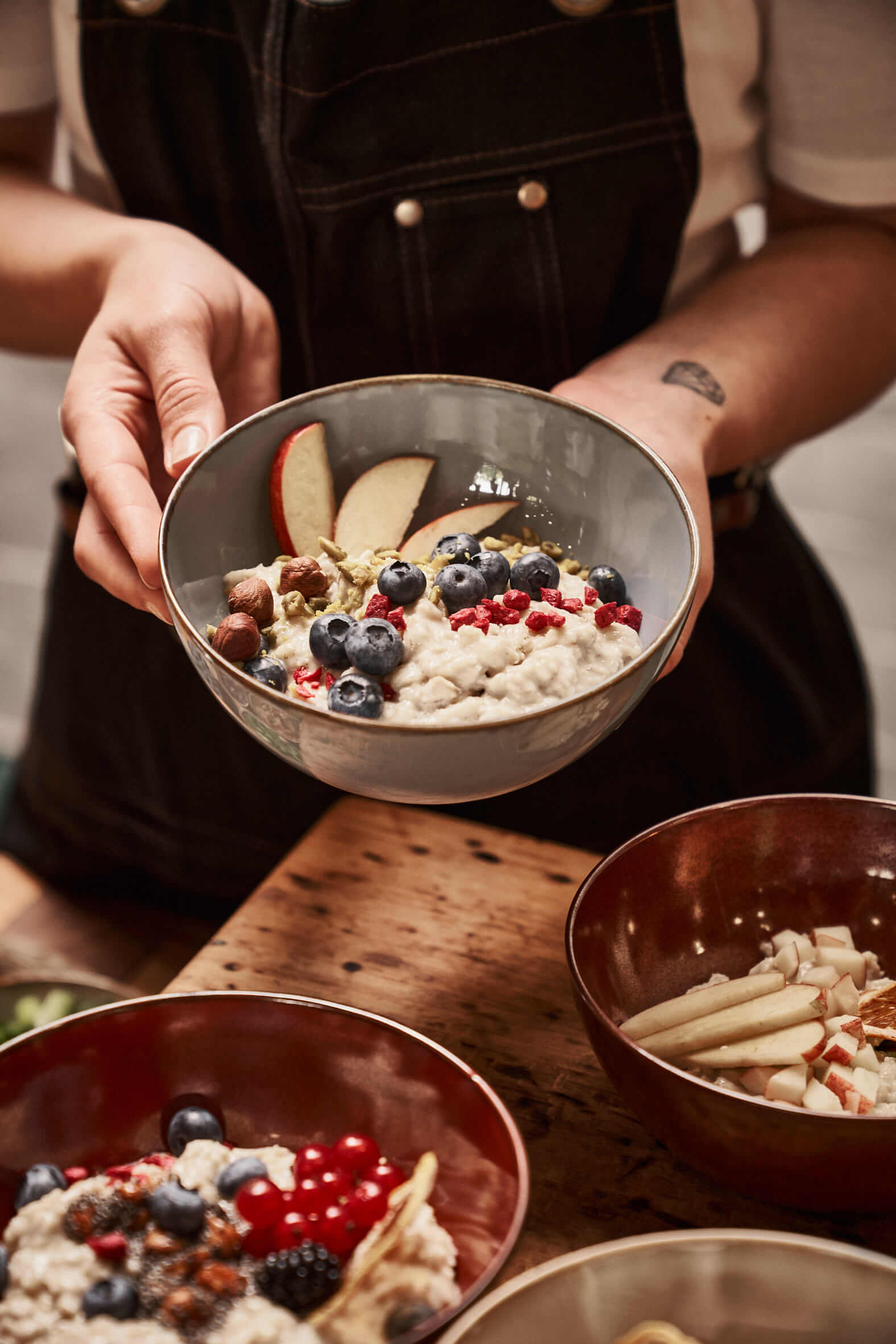 A woman holds a bowl filled with porridge, berries, nuts and fruits in front of her. On the table below are more porridge bowls.