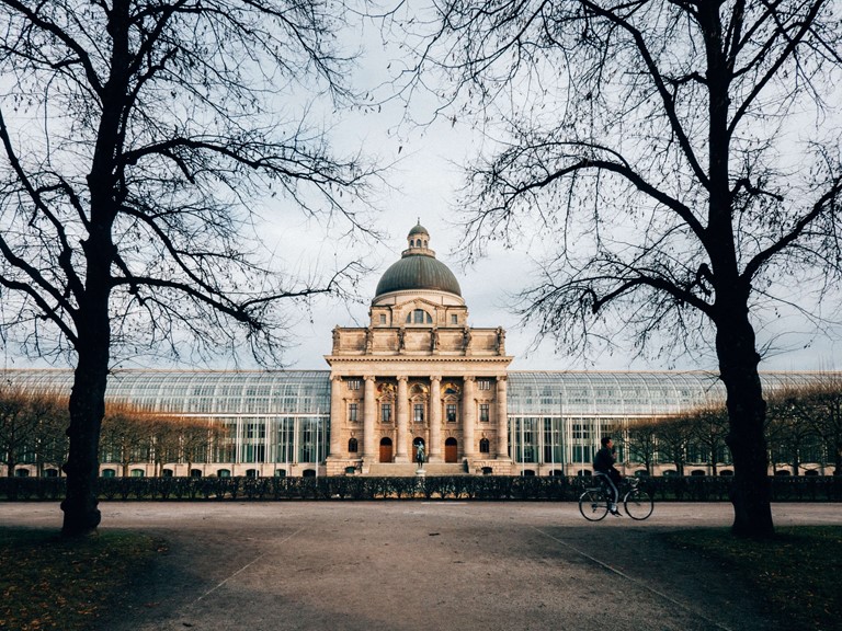 A historic building with a dome and glass extensions on the right and left in a park, a cyclist moves away in front of it