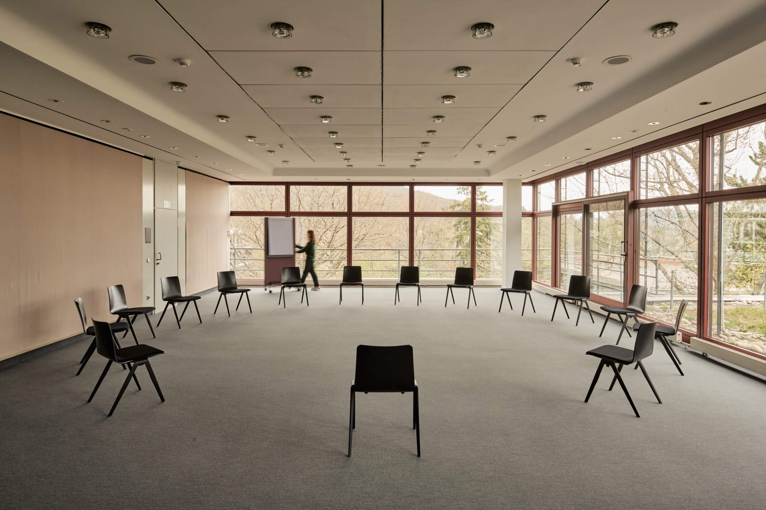 A conference room with floor-to-ceiling windows. Chairs are arranged in a circle in the centre of the room. In the background, a woman is pushing a flipchart.