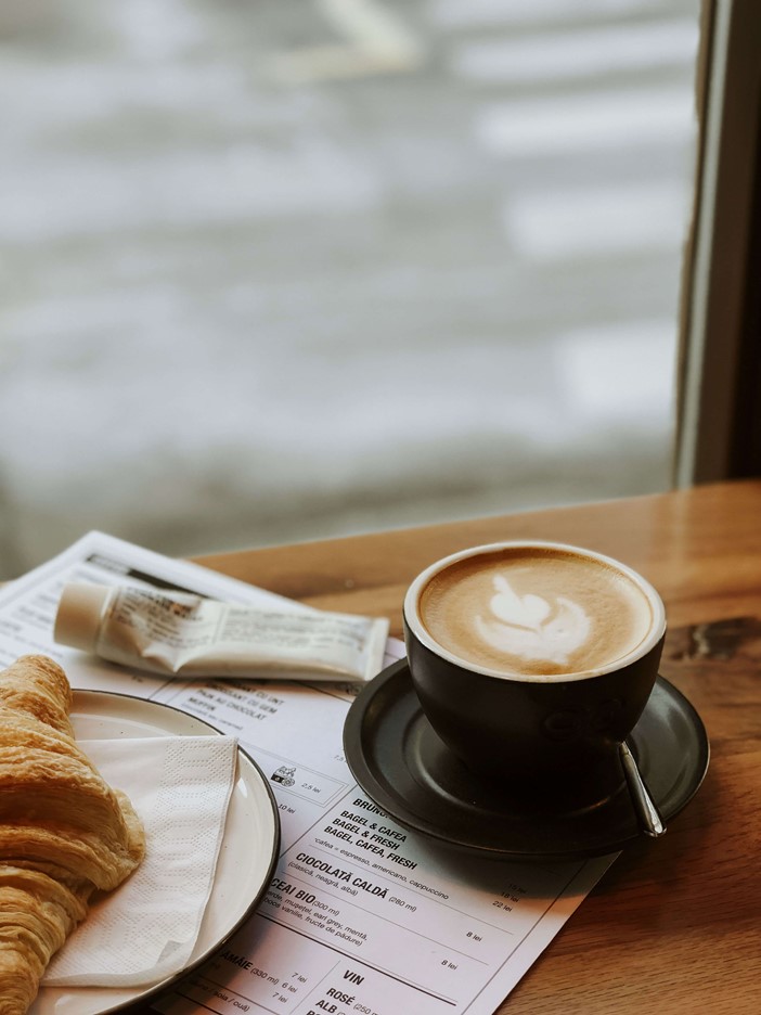 Close-up of a breakfast on a wooden table with a view to the outside. On the right a coffee and on the left a croissant on a plate, below a menu.