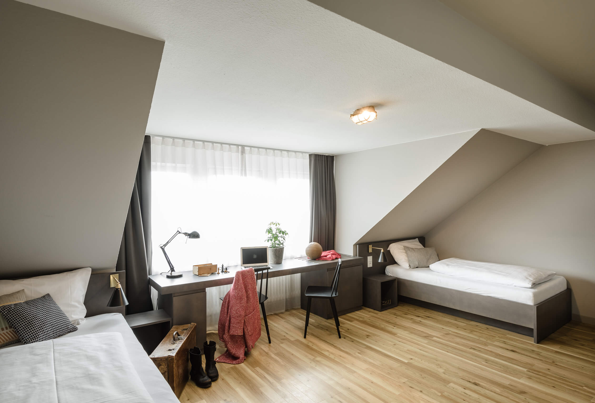 Blick in ein Zimmer mit hellem Holzboden. Rechts und links stehen Einzelbetten, mittig befindet sich ein breites Fenster mit Vorhängen, davor ein Schreibtisch mit Accessoires.