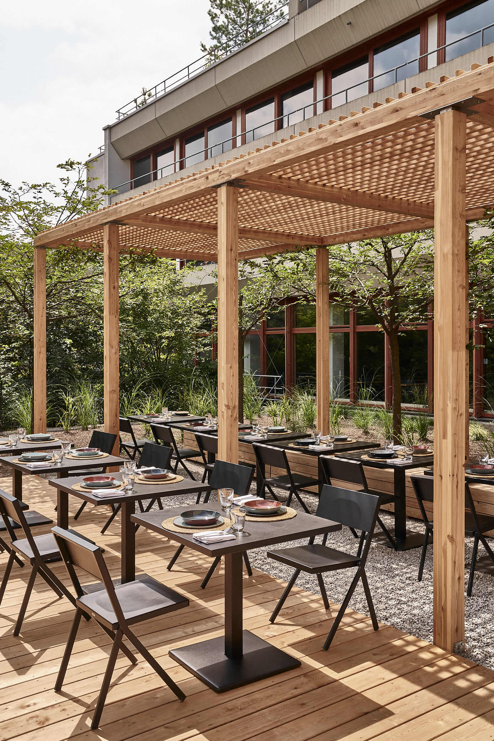Covered black tables and chairs, partly on light wooden planks and partly in the half-shade of a wooden pergola. 