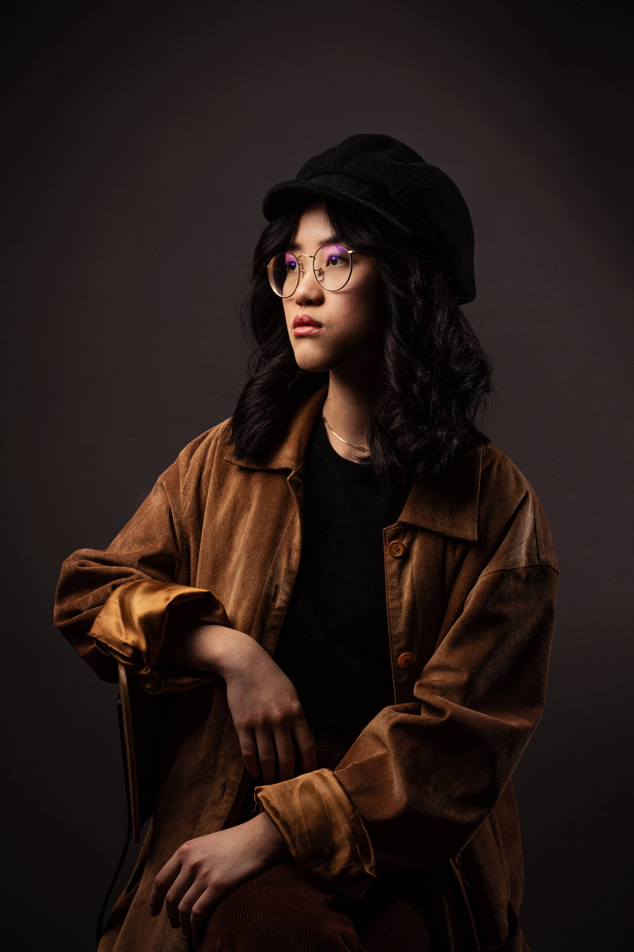 Eine asiatisch anmutende Frau mit eingedrehten schwarzen Haaren, großer, runder Brille und schwarzem Hut posiert im Stil alter Meister vor einem dunklen Hintergrund. Sie trägt eine cognacfarbene Oversize-Jacke.